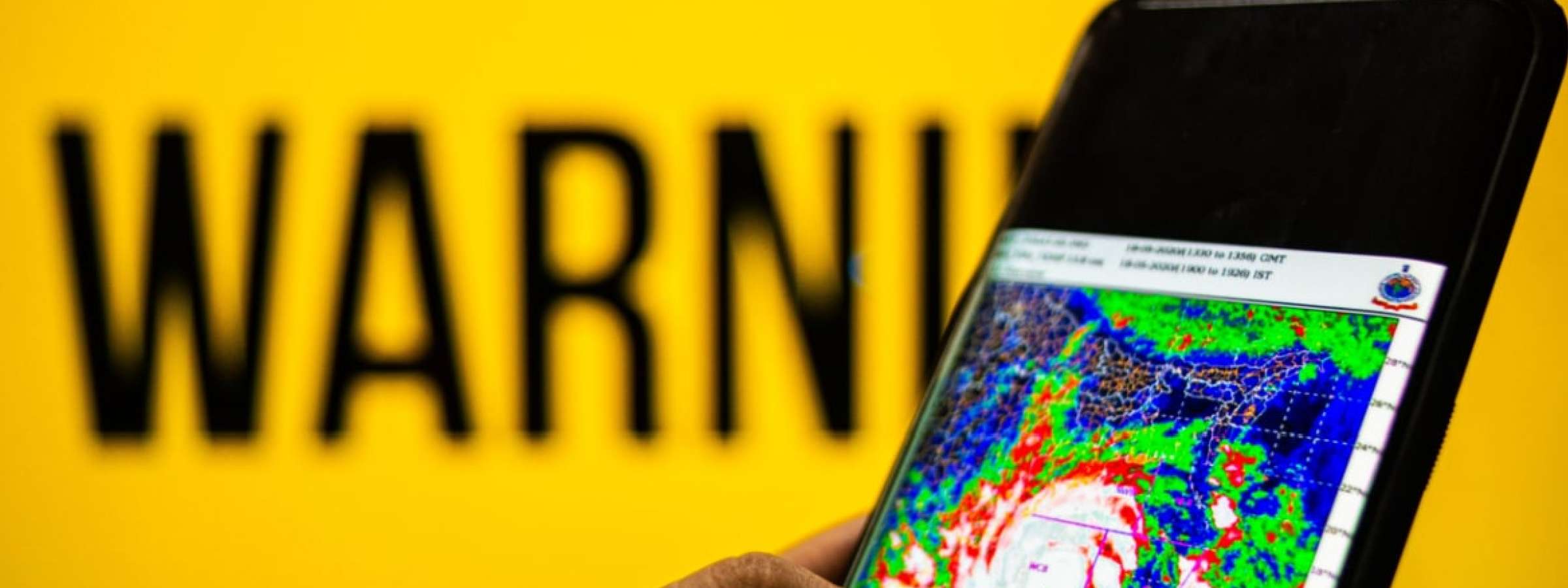 Photograph of a mobile phone with cyclone moving towards the coast shown. The background is yellow with warning sign.